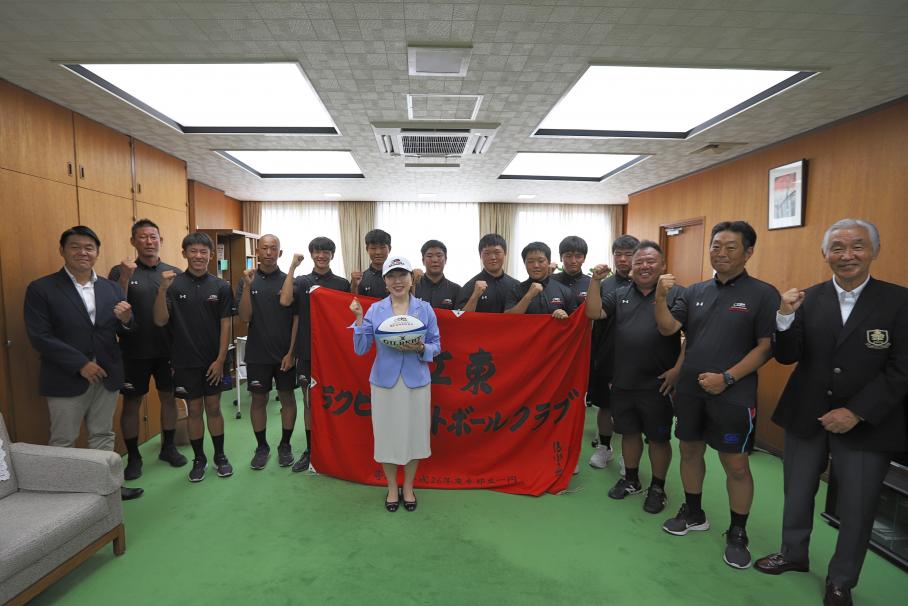「江東ラグビークラブ」と書かれた赤い旗を手に持つ中学生選手・コーチらと、ラグビーボールと手に持ち中央に立つ木村区長