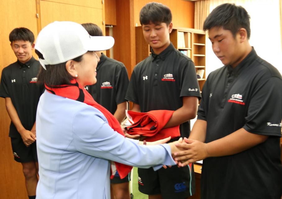 赤い旗を首にかけ、白いキャップを頭にかぶり、中学生選手と握手する木村区長
