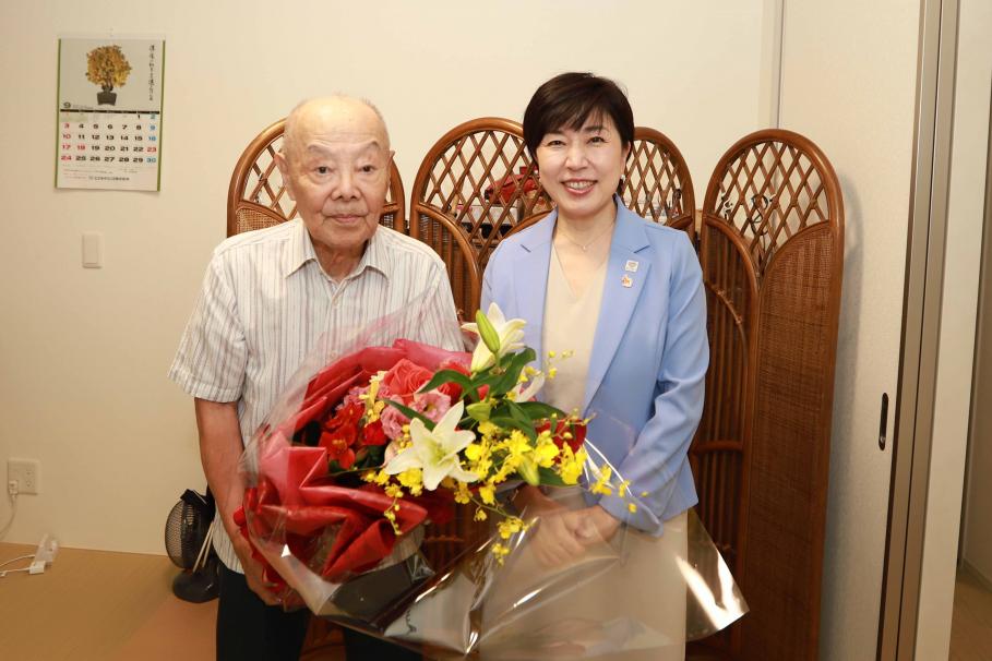 ユリや黄色い小さな花などでつくられた大きな花束を手に木村区長と写る岡田さん。白地にストライプの柄の半そでワイシャツを着ている。
