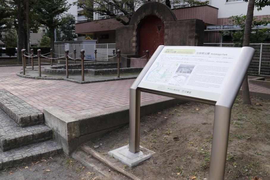 元加賀公園の写真。右手前には元加賀公園について記載された、白い説明版があり、奥には赤茶のレンガでできたアーチ状の壁泉がある。