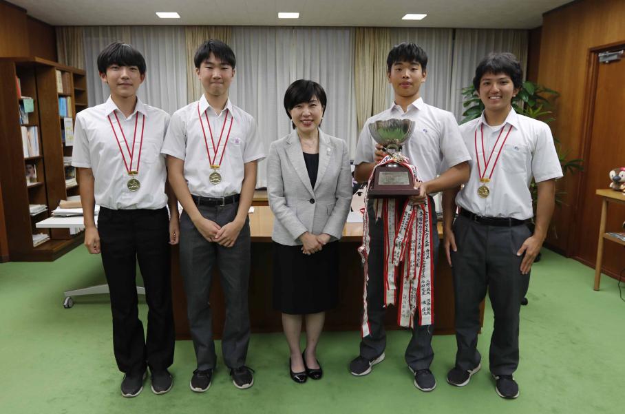 区長室で首から金メダルをさげて木村区長を中央に挟み、写真に写る選手4名。右から2番目の塚本さんは手にトロフィーを持っている。