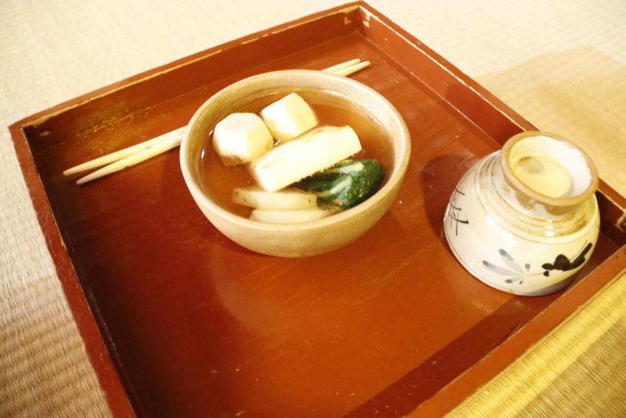 正方形の赤茶のお盆には雑煮と箸、茶碗が乗る。雑煮は透明の茶色い汁に里芋、小松菜、大根等が浮いたもの。