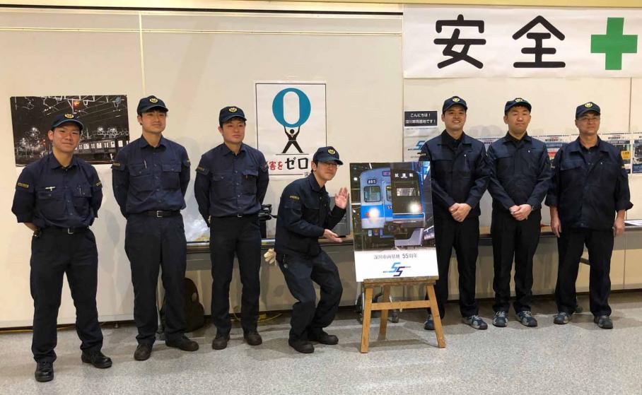東西線車両を正面から撮影した55周年記念ポスターを囲んで立つ、東京メトロの職員7名。濃紺の作業着にキャップをかぶっている。
