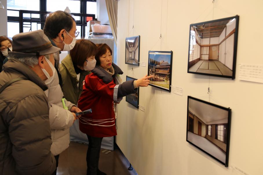 壁にかけられた旧渋沢家住宅外観の写真などを指さして解説する文化観光ガイドと、身を乗り出して写真を見る男女の利用客4名