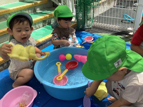 1歳児がテラスでたらいに入った水をひしゃくやじょうろで流して遊んでいる様子の写真