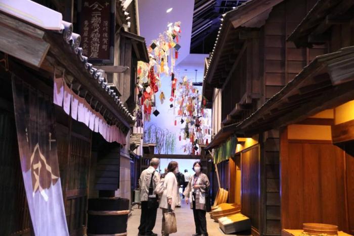 江戸時代の店等を再現した建物が並ぶ奥には、高さ約8メートルの笹竹2本が立っており、七夕飾りがつけられている。手前には職員と来館者