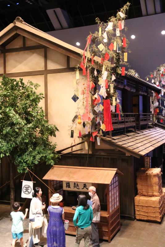 江戸の天ぷら屋の屋台の前で江戸のまちについて職員から説明を受ける家族連れ。奥には高さ８メートルの竹が。