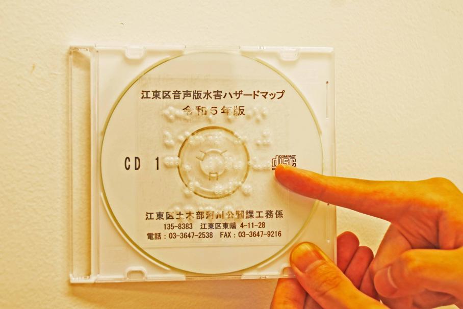 音声版水害ハザードマップCDの写真。透明なCDケースの表面には点字シートが貼られている。CDの色は白。