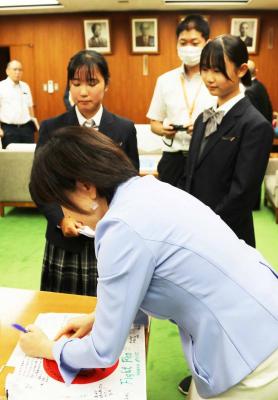 区長室の机で選手が持参した日本国旗に青いペンで応援メッセージを書く区長。区長の横には選手2名と区職員。