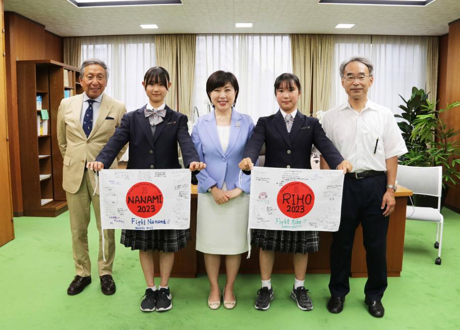 区長室の机の前で写真に写る区長、中学生セーリング選手2名、コーチ、校長。選手はそれぞれ手に応援メッセージ入りの日本国旗を持つ。。