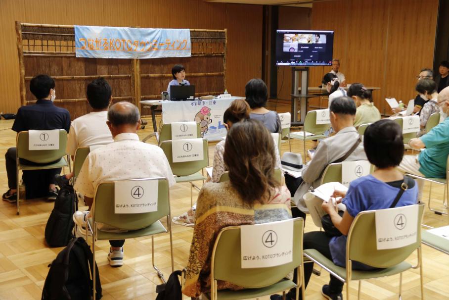 椅子に座る参加者を前に腰かけて6つの公約について説明する木村区長。区長の隣のサイネージにはオンライン参加の5名の顔も映されている