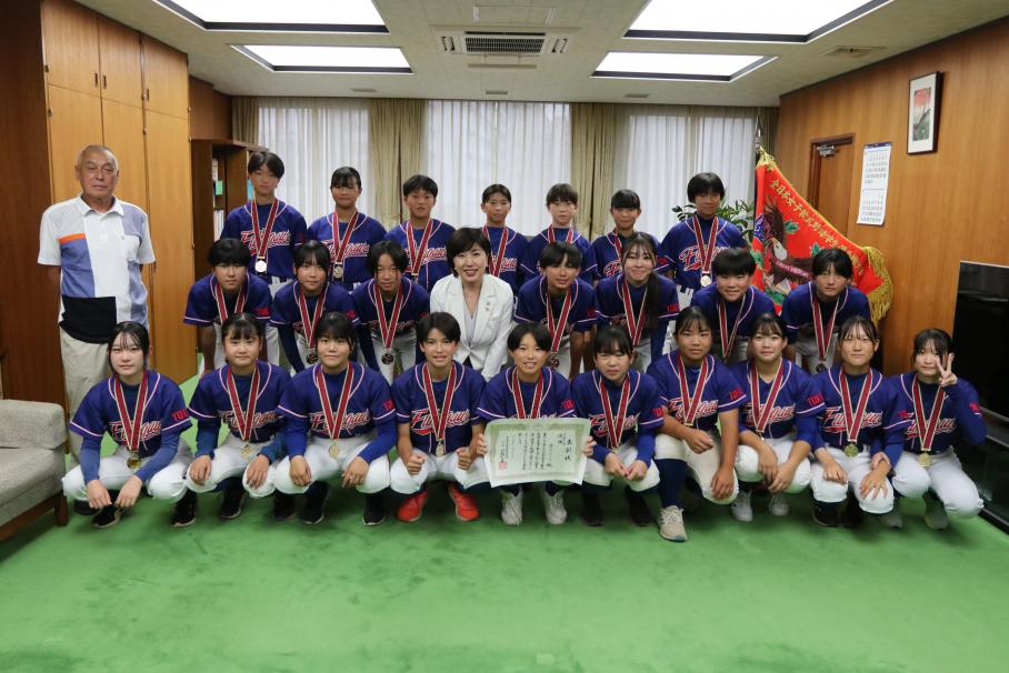 白いジャケットの木村区長を中央に囲み、青いユニホームと金メダルで集合する選手24人。左側にはコーチ、一番後ろ右には赤い優勝旗。