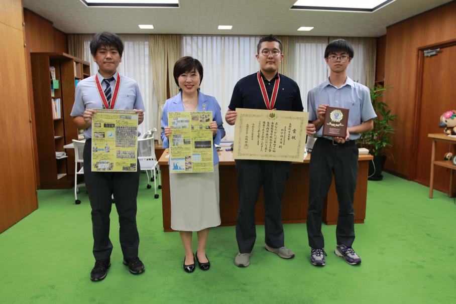 木村区長を囲んで立つ、科学研究部の城さんたち3名。3名のうち2名は首からメダルを下げ賞状・ポスターを持ち、1名は盾を持っている。