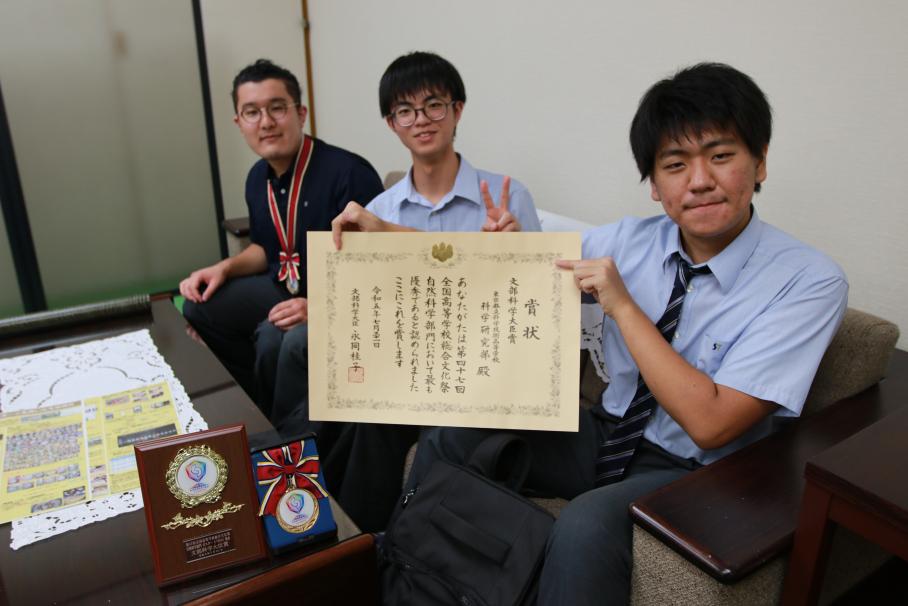 ソファに座りカメラ目線で写る科学技術高校の部員3名。一番手前の生徒は賞状を掲げ、中央の生徒はピースをしている。机には盾が。