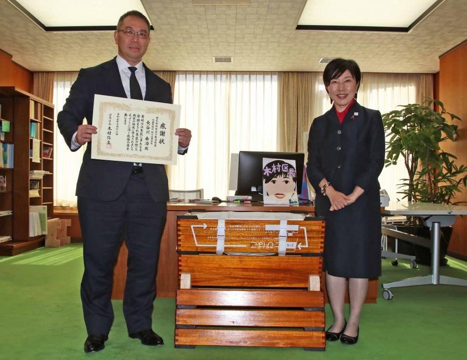 黒いスーツに身を包み、手には感謝状を持つ長谷川代表取締役。中央にトランスフォームとび箱を置き、右側には紺のスーツを着用した区長