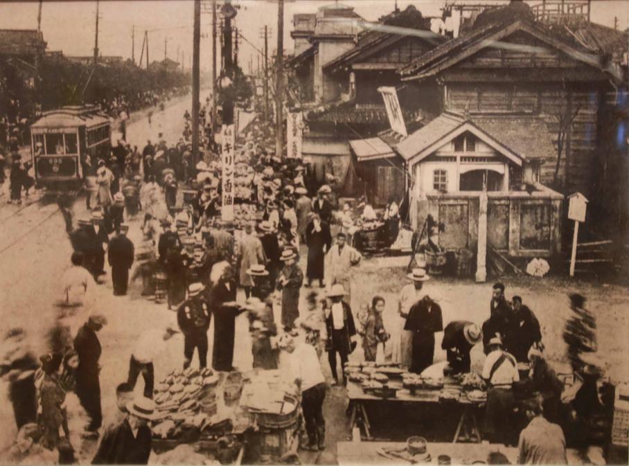黒江町の写真。左側に永代通りが走るが幅は狭い。木造の家屋が立ち並び、手前では何かの叩き売りが行われ、多くの人だかりができている。