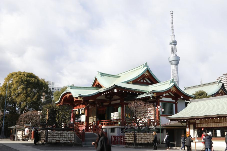 青磁の色の瓦屋根に赤い柱が特徴的な亀戸天神社。手前には参拝者が3名程おり、亀戸天神社の後ろにはスカイツリーが見える。